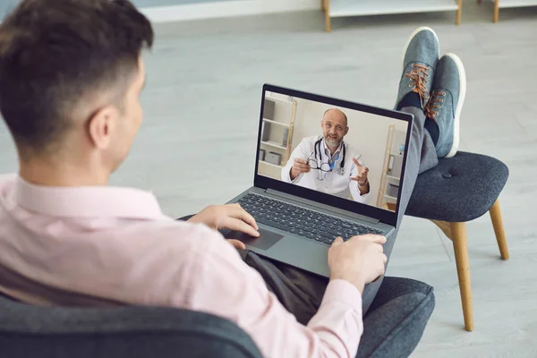 Dottore online. Un uomo seduto su una sedia ha una videochiamata che aiuta un medico online maturo con un computer portatile nel suo appartamento . — Foto Stock