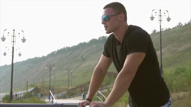 Young sporty man in sunglasses having a rest on seafront and look at the water — Stock Video