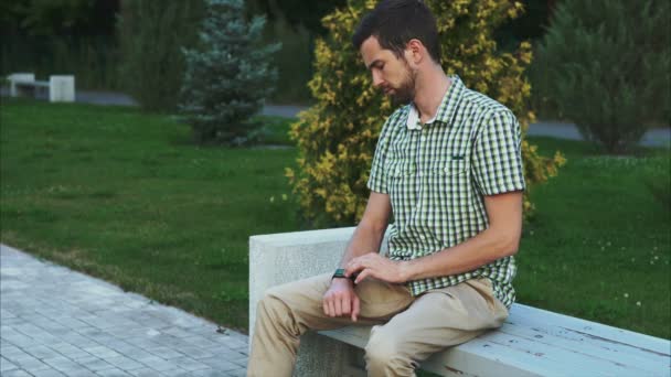 Hombre vestido casual sentarse en el banco en el parque y utilizar su reloj inteligente moderno — Vídeos de Stock