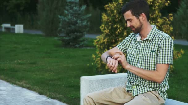 Casual vestido homem sentar no banco no parque e usar o seu relógio inteligente moderno — Vídeo de Stock