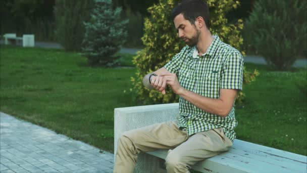 Mladý muž, zatímco sedí na lavičce v parku pomocí hodinek smartwatch — Stock video
