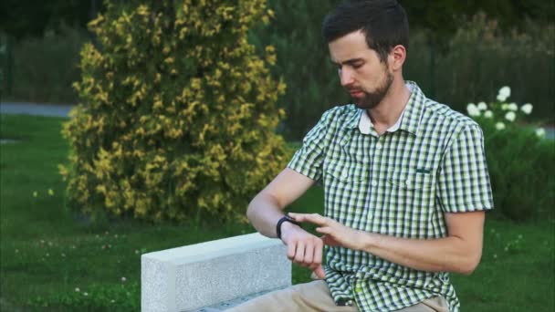 Joven usando smartwatch mientras está sentado en el parque — Vídeo de stock