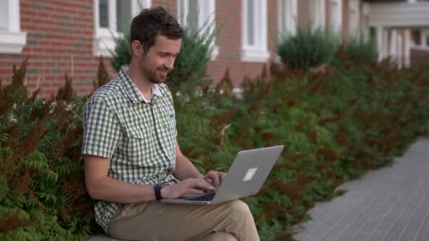 Случайно одетый мужчина-фрилансер сидит на скамейке и пользуется своим ноутбуком — стоковое видео