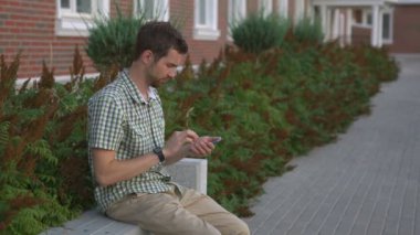 Casual giyimli adam bankta oturmak ve onun akıllı izlemek ve smartphone kullanın