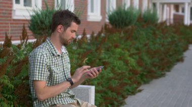 Casual giyimli adam bankta oturmak ve onun smartwatch ve smartphone kullanın