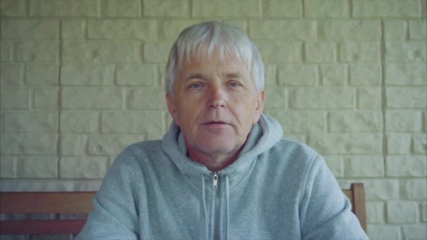 Porträt eines älteren Mannes mit grauen Haaren vor dem Haus, der auf der Bank sitzt — Stockvideo