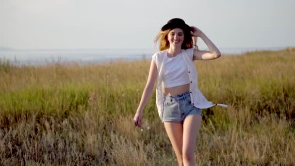 Young beautiful girl model in hat runnig and dancing on meadow near the river — Stock Video