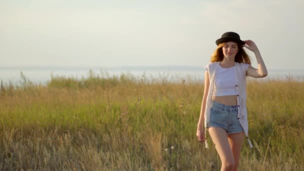 Молодая красивая девушка модель в шляпе ходить и позировать на лугу у реки — стоковое видео