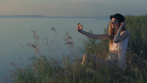 Νεαρό κορίτσι κάνει selfie που κάθεται στην άκρη ενός γκρεμού, κοντά στον ποταμό — Αρχείο Βίντεο