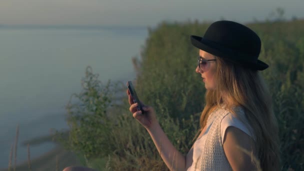 Young girl use her smartphone sitting on the edge of a cliff near the river — Stock Video