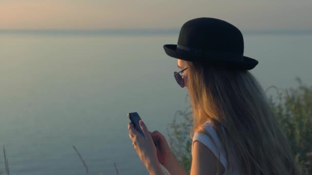 Chica joven utilizar su teléfono inteligente sentado en el borde de un acantilado cerca del río — Vídeo de stock