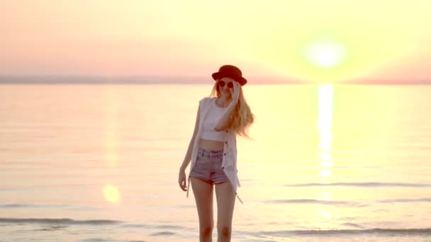 Bonita joven rubia con sombrero posando sobre el fondo del océano y la puesta de sol dorada — Vídeo de stock