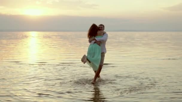 Joven hombre feliz recoger y girar a su novia en la playa en la puesta de sol — Vídeos de Stock