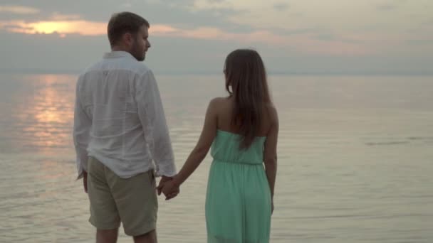 Feliz jovem casal na praia no pôr do sol eles dão as mãos e olhar uns para os outros — Vídeo de Stock