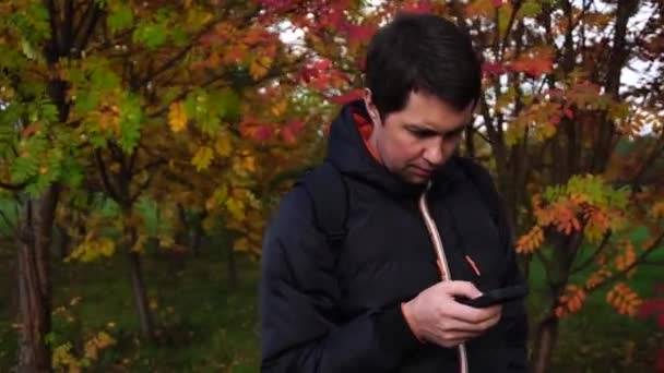 年轻人穿黑夹克在秋天公园使用他的智能手机 — 图库视频影像
