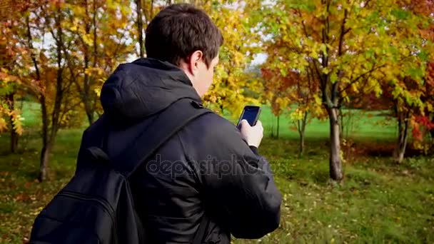 在使用他的智能手机的秋天公园散步的黑色外套的年轻人 — 图库视频影像