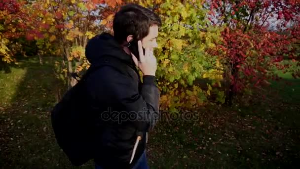 Jonge man in zwarte jas, wandelen in de herfst park praten aan de telefoon — Stockvideo