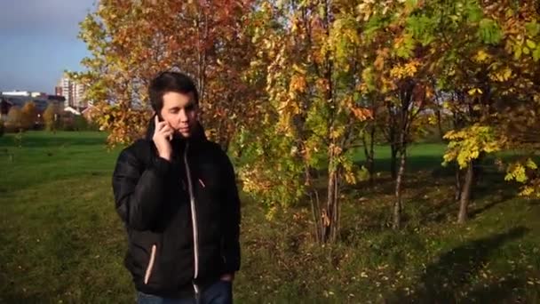 Молодой человек в черной куртке, гуляющий в осеннем парке и разговаривающий по телефону — стоковое видео