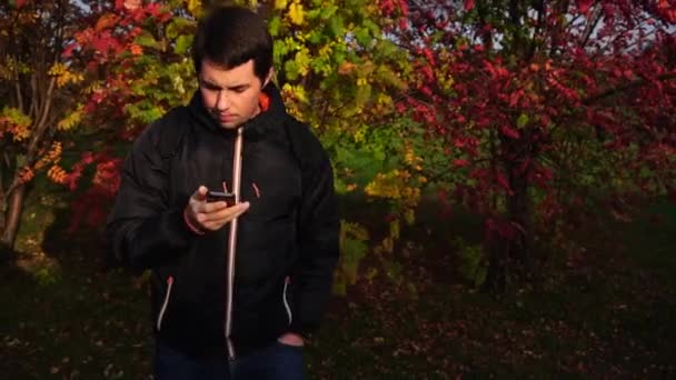 Junger Mann in schwarzer Jacke im Herbstpark mit Smartphone — Stockvideo