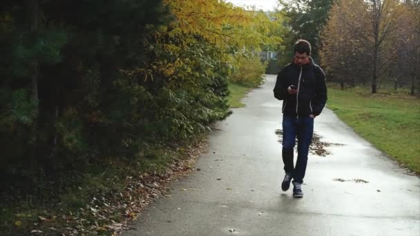 Молодой человек в черной куртке гуляет по осеннему парку со своим смартфоном — стоковое видео