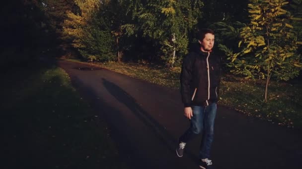 Молодой спокойный человек в черной куртке, прогулка в осеннем парке драматический свет — стоковое видео