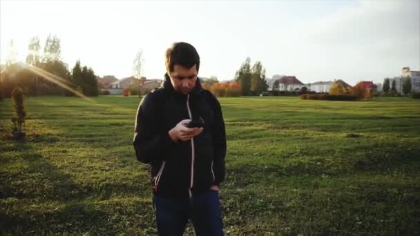 Hombre joven con chaqueta negra navegar por Internet usando su teléfono inteligente al aire libre — Vídeo de stock