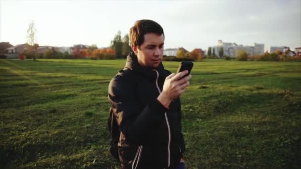 Jovem de casaco preto navegar na internet usando seu smartphone ao ar livre — Vídeo de Stock