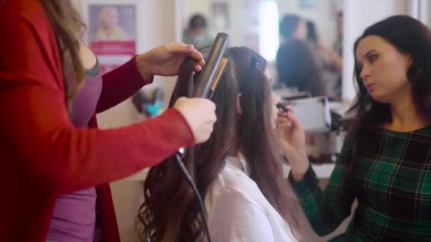 Hairstylist making hair-do with hair iron while make up artist applying cosmetic — Stock Video
