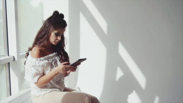 Joven guapa morena usando su tableta sobre fondo blanco cerca de la ventana — Vídeo de stock