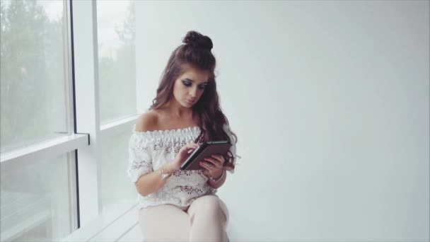 Красивая брюнетка с помощью планшета на белом фоне возле окна — стоковое видео