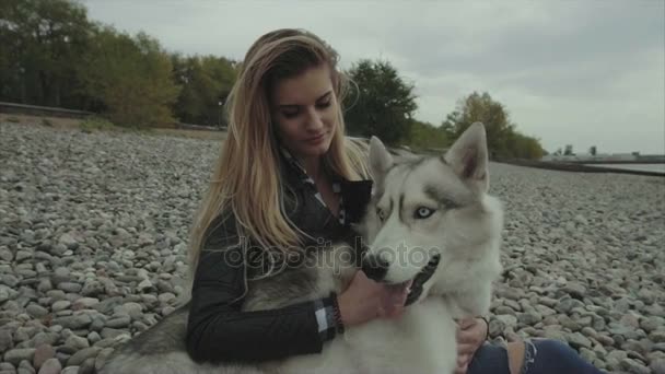 Joven chica hermosa con perro husky siberiano sentado en la orilla del río — Vídeo de stock