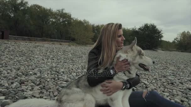 Νεαρή όμορφη γυναίκα με Σιβηρικό γεροδεμένο σκυλί κάθεται στην όχθη του ποταμού — Αρχείο Βίντεο