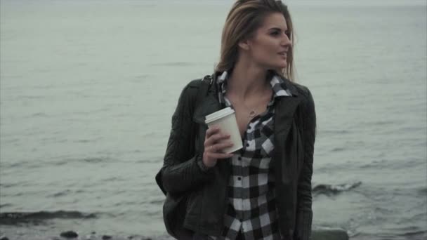 Hermosa joven rubia sosteniendo una taza de café en sus manos cerca del mar — Vídeo de stock