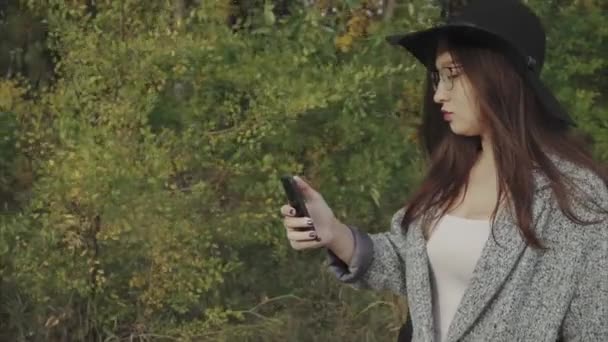 Joven mujer bonita en sombrero negro y gafas de escribir sms a su novio al aire libre — Vídeos de Stock