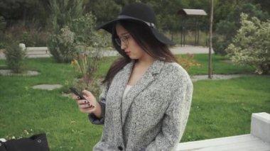 Genç ve güzel kadın siyah şapka ve gözlük şehir parkında smartphone kullanarak