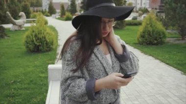 Genç ve güzel kadın siyah şapka ve gözlük şehir parkında smartphone kullanarak