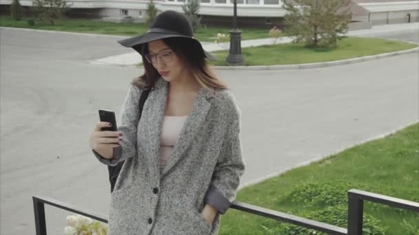 Mooie vrouw in zwarte hoed en bril texting op smartphone buiten — Stockvideo