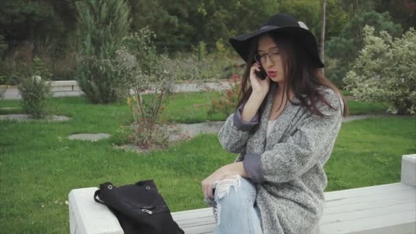 Jovem mulher bonita em chapéu preto e óculos falando no telefone ao ar livre em um parque — Vídeo de Stock