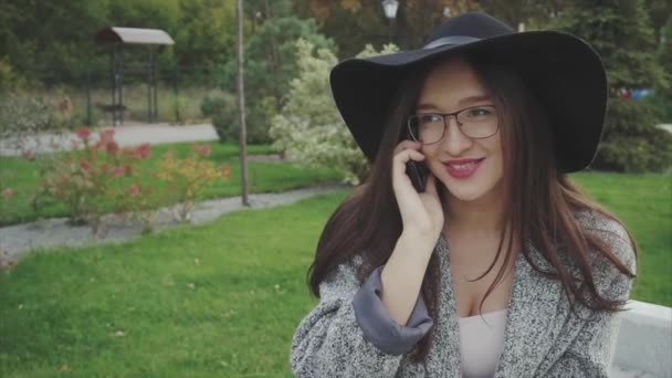 Jovem mulher de chapéu preto e óculos falando no telefone senta-se no banco no parque — Vídeo de Stock