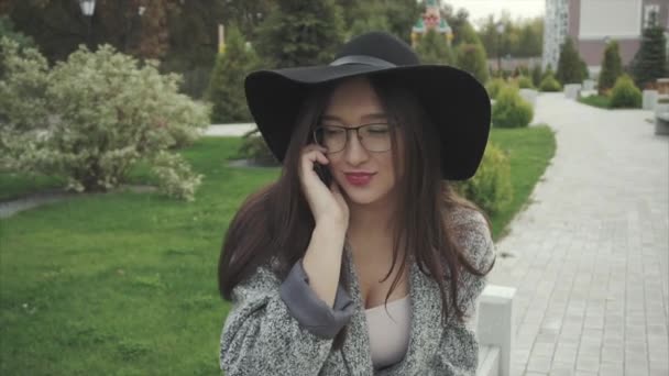 Mladá žena v černém klobouku a brýle mluvit na mobilním telefonu v městském parku — Stock video