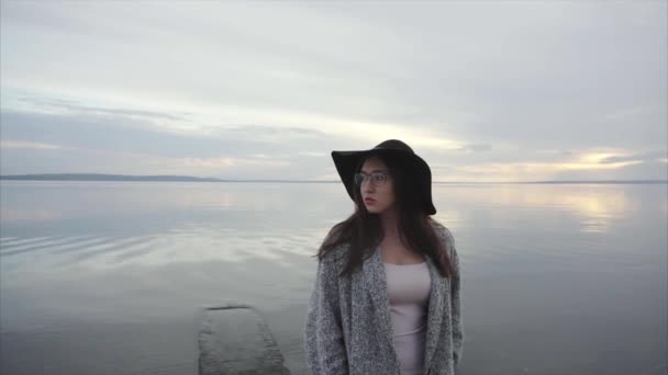 Mujer joven con sombrero negro y gafas mirando alrededor de la costa al atardecer — Vídeo de stock