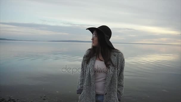 Młoda kobieta w czarny kapelusz i okulary w pobliżu morza na zachód słońca — Wideo stockowe
