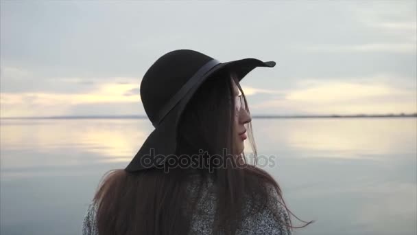 Mujer joven con sombrero negro y gafas cerca del mar al atardecer — Vídeos de Stock