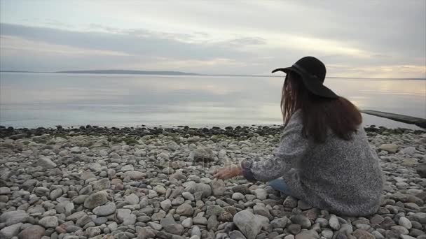 Mujer joven sentada en la playa de guijarros arrojando piedras al agua — Vídeos de Stock