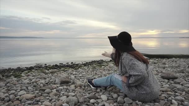 Suya taş atan çakıl plajı üzerinde oturan genç kadın — Stok video