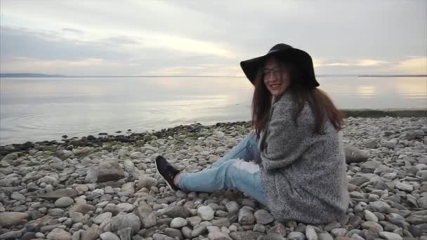 Молодая красивая женщина в черной шляпе и очках, сидящая у моря на закате — стоковое видео