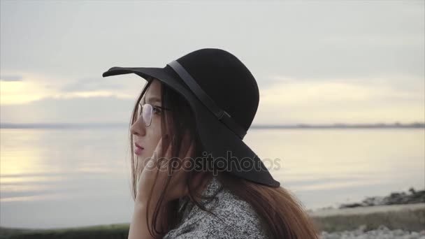 Joven mujer bastante sonriente en sombrero negro y gafas sentadas cerca del mar al atardecer — Vídeos de Stock