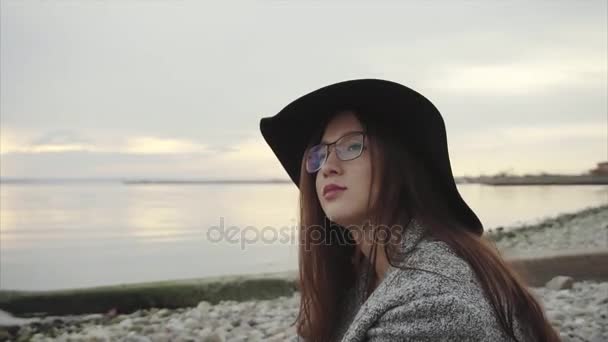 Mulher bonita bonita wistful no chapéu preto e nos óculos que sentam perto do por do sol do mar — Vídeo de Stock