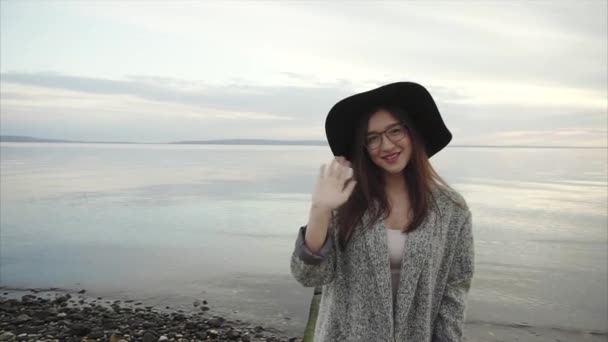 Joven mujer bonita en sombrero negro y gafas agitando una mano cerca del mar al atardecer — Vídeos de Stock