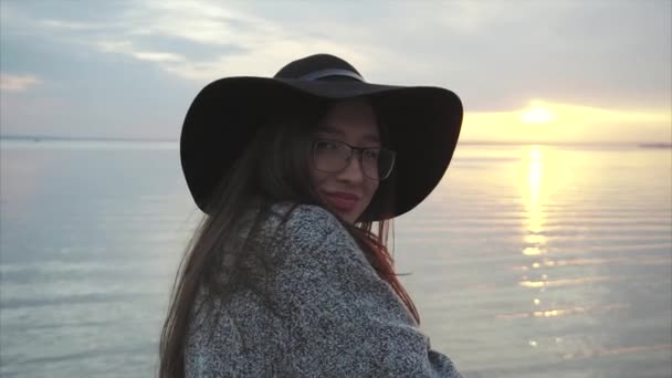 Молода досить усміхнена жінка в чорному капелюсі і окулярах біля моря на заході сонця — стокове відео